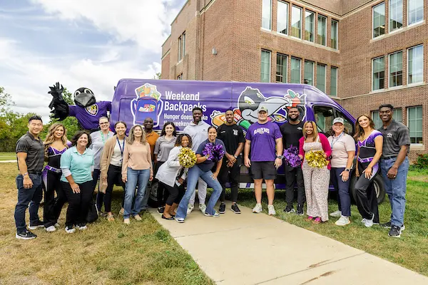 Ravens players, mascot, and cheerleaders, Weekend Backpacks staff, and school staff standing in front of the Weekend Backpacks van at an elementary school