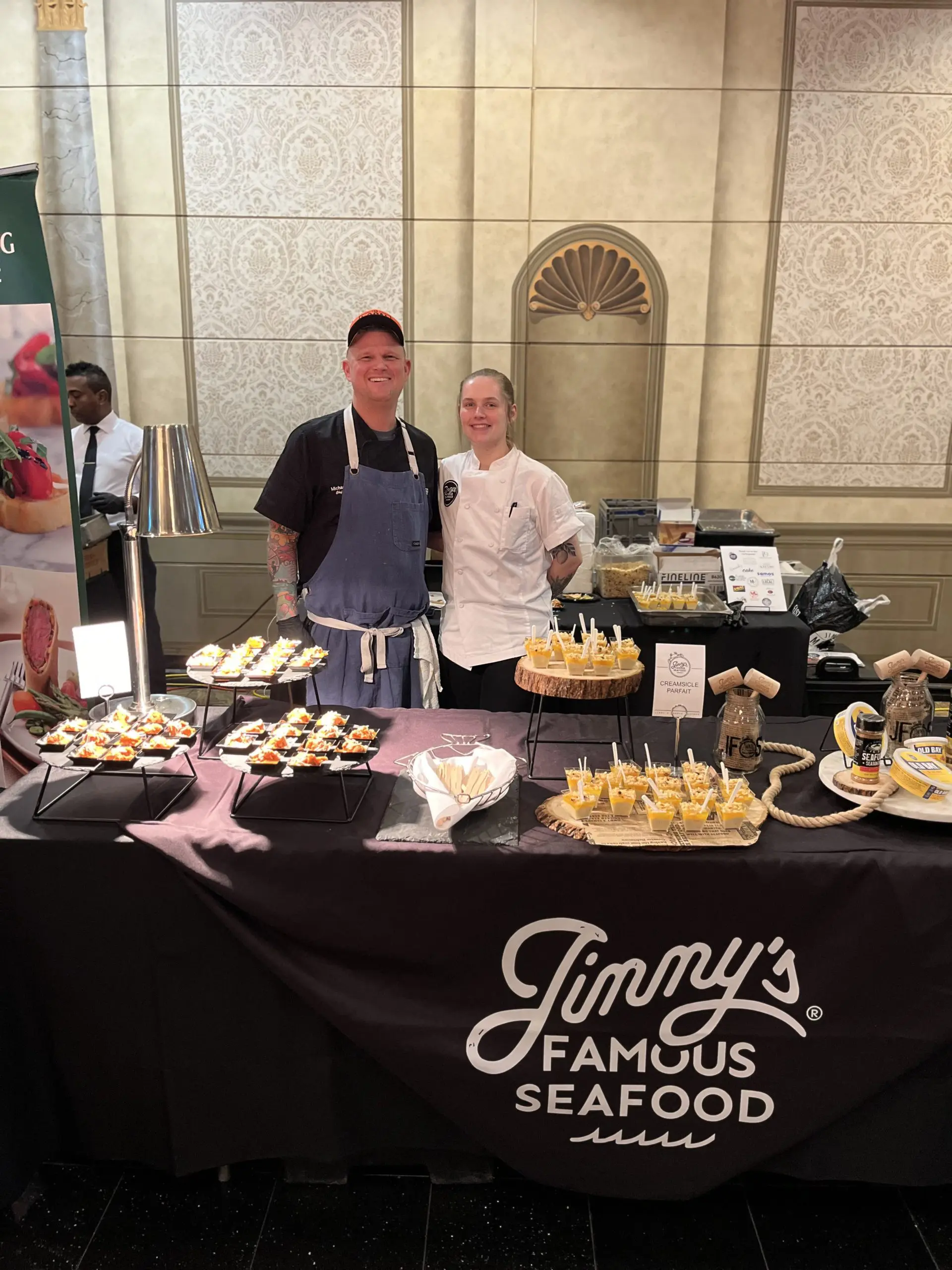 Two chefs posed behind a table of their food samples with a sign saying "Jimmy's Famous Seafood."