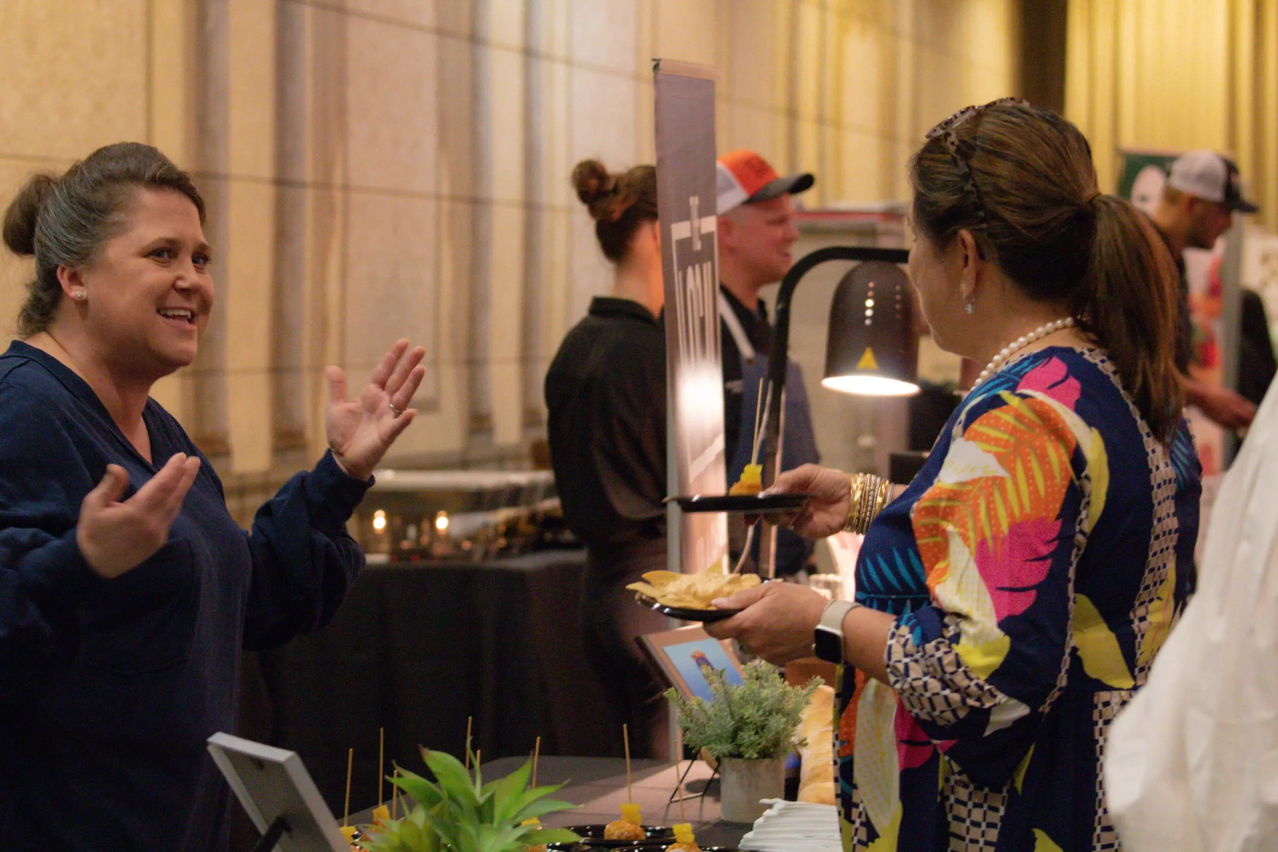 A woman talking to a chef at a table of food samples.