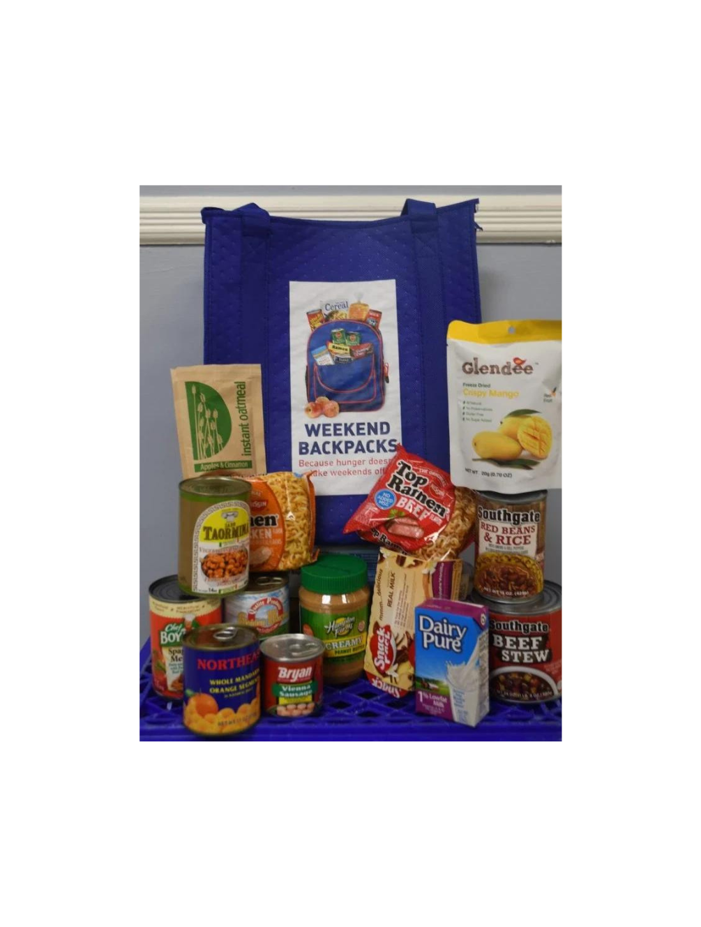 Various non-perishable foods including peanut butter, ramen noodles, milk, oatmeal, a can of fruit, and prepared meals in front of a tote bag with the Weekend Backpacks logo.