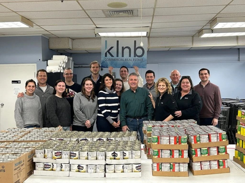 A group of people posed behind a table with cases of food holding a sign that says "klnb commercial real estate."