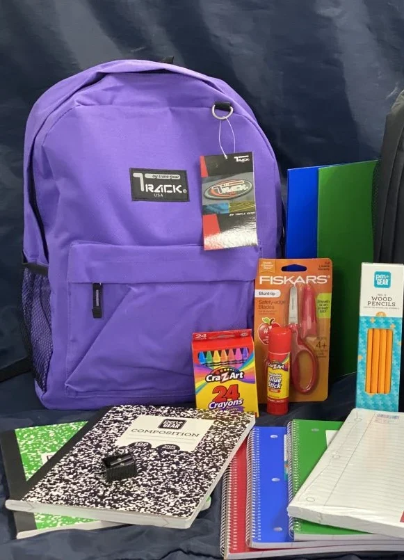 A backpack with school supplies including notebooks, crayons, pencils, scissors, and paper laid out in front of it.