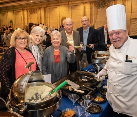 A chef and five patrons standing at his table smiling.