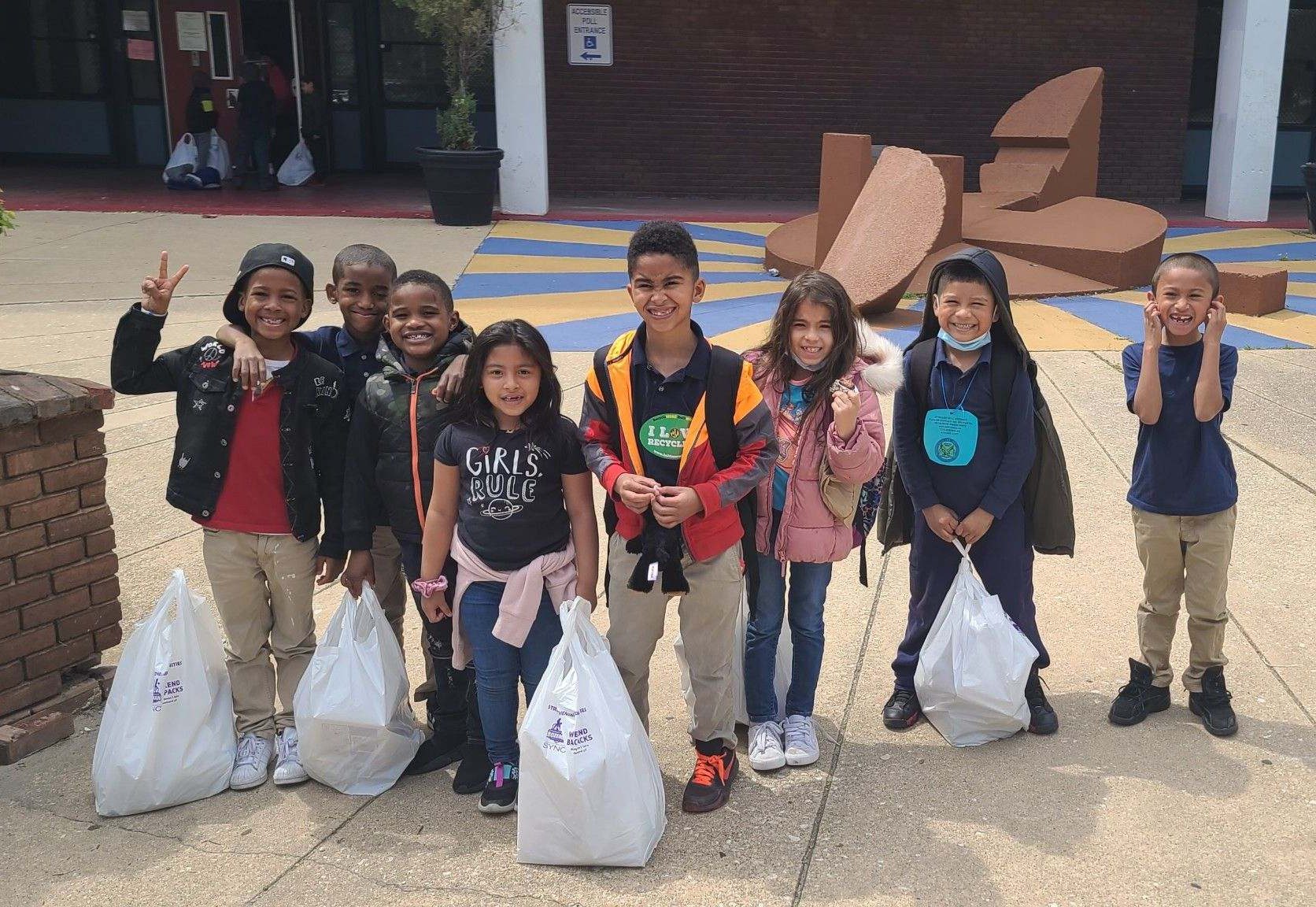 Eight young students standing in front of their school smiling and holding plastic bags of food.